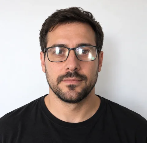 AI-generated photo of a man with a beard wearing dark rectangular eyeglasses with light reflecting on the lenses, in front of a white wall